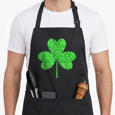 Irish shamrock st patrick day aprons - Minimalist green shamrock with three heart-shaped leaves and stem centered — shown on a black cotton canvas apron against a white background