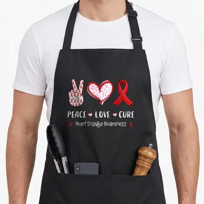 Peace heart disease awareness apron - Red and white cartoon peace sign, heart, and ribbon with text PEACE LOVE CURE — shown on a black cotton canvas apron against a white background