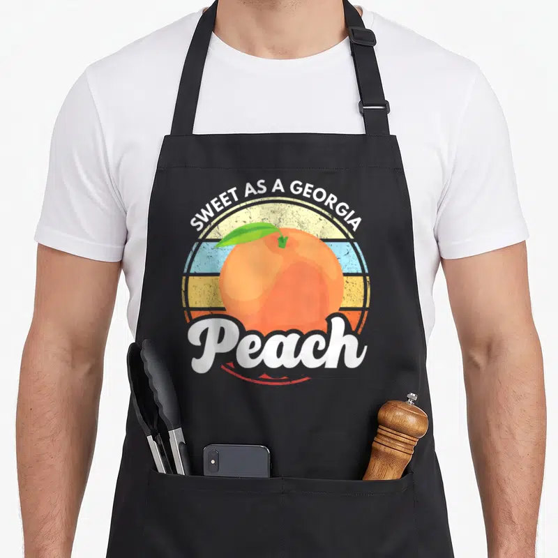 Belle southern aprons - Orange peach with green leaf over multicolored striped circle, white text SWEET AS A GEORGIA — shown on a black cotton canvas apron against a white background