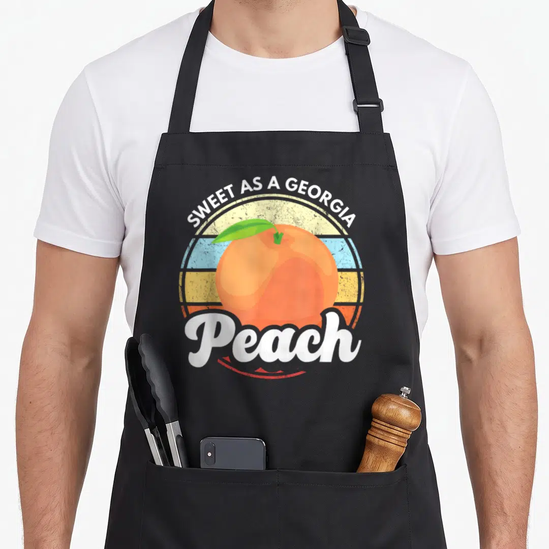 Belle southern aprons - Orange peach with green leaf over multicolored striped circle, white text SWEET AS A GEORGIA — shown on a black cotton canvas apron against a white background