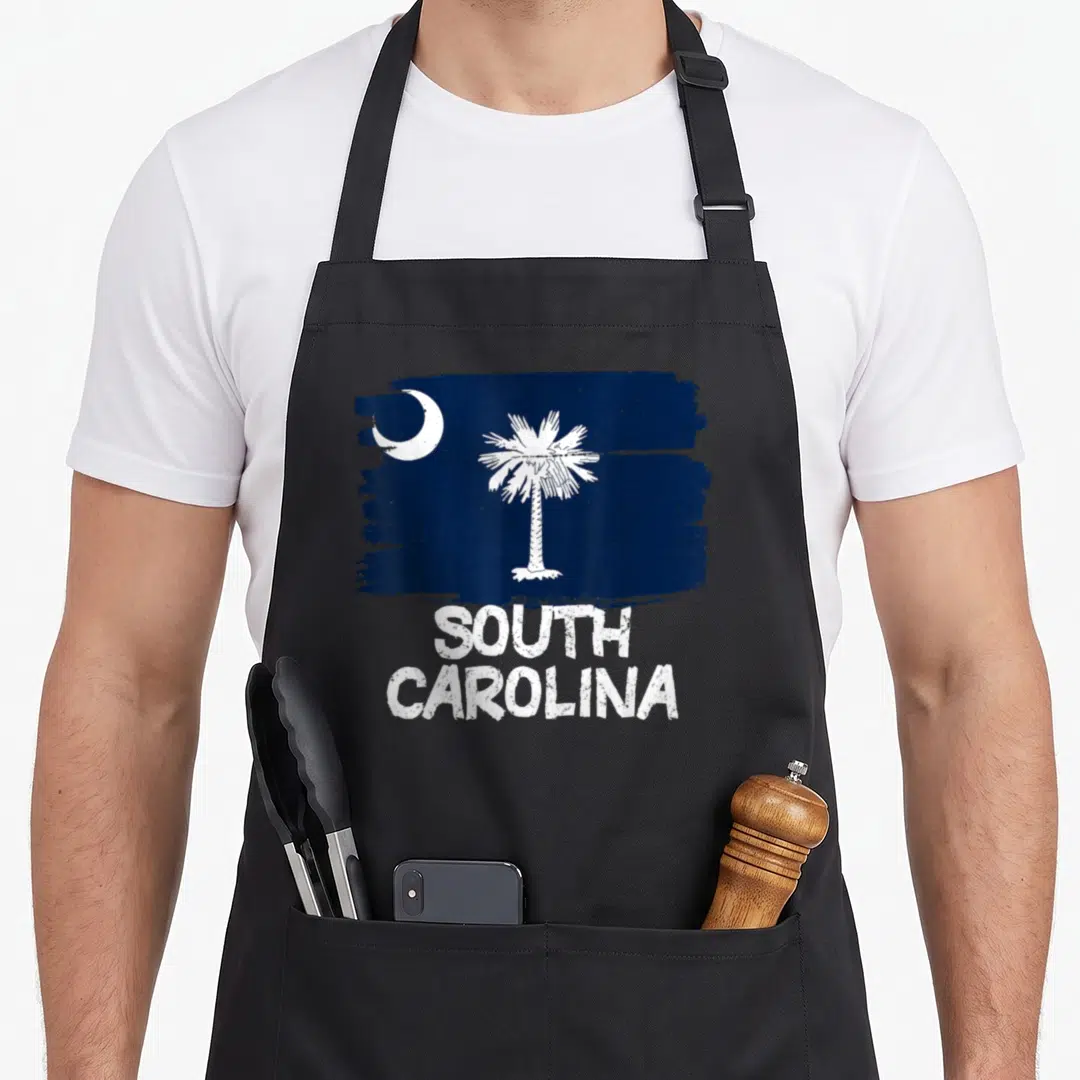 Flag carolina apron - Distressed dark blue flag with white moon, palm tree, and stacked SOUTH CAROLINA text — shown on a black cotton canvas apron against a white background