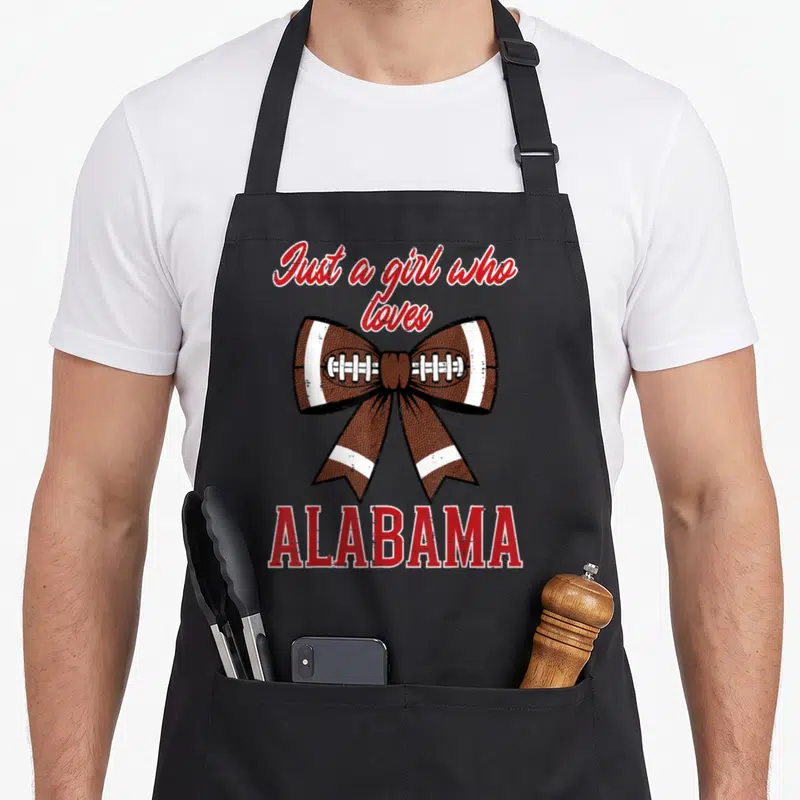 Funny youth alabama apron - Brown football texture bow with white laces and red Just a girl who loves ALABAMA text — shown on a black cotton canvas apron against a white background