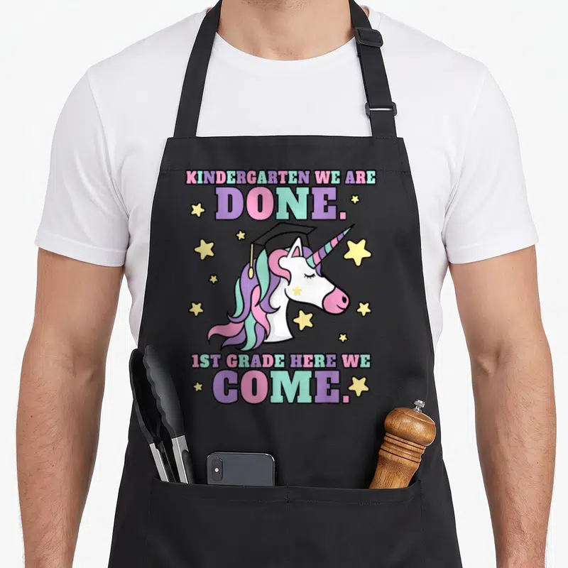 Graduation magical unicorn aprons - Cartoon unicorn with graduation cap surrounded by stars and stacked text in purple — shown on a black cotton canvas apron against a white background