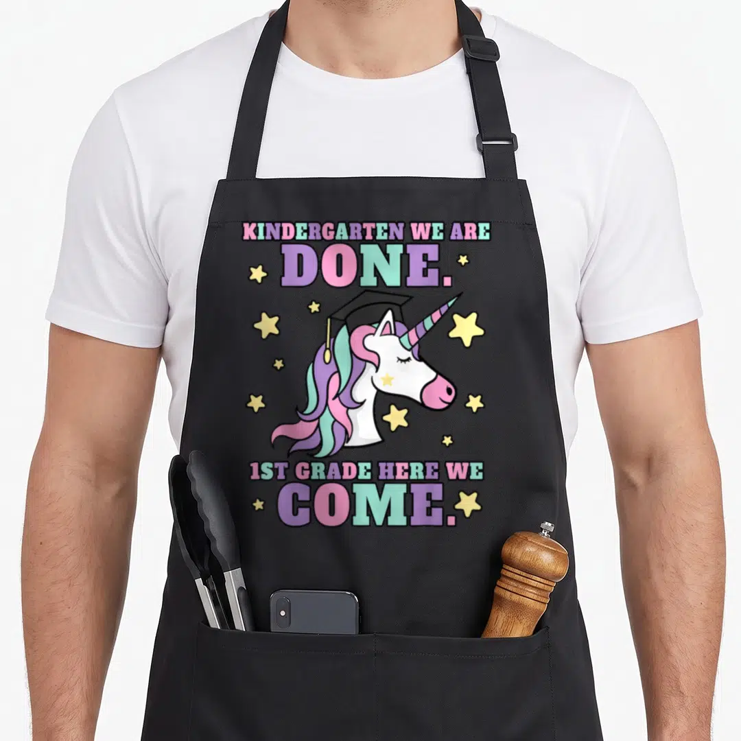 Graduation magical unicorn aprons - Cartoon unicorn with graduation cap surrounded by stars and stacked text in purple — shown on a black cotton canvas apron against a white background
