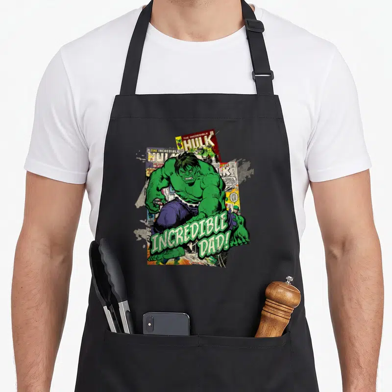 Cute incredible father day apron - Hulk illustration over three comic covers with INCREDIBLE DAD! text in green letters — shown on a black cotton canvas apron against a white background