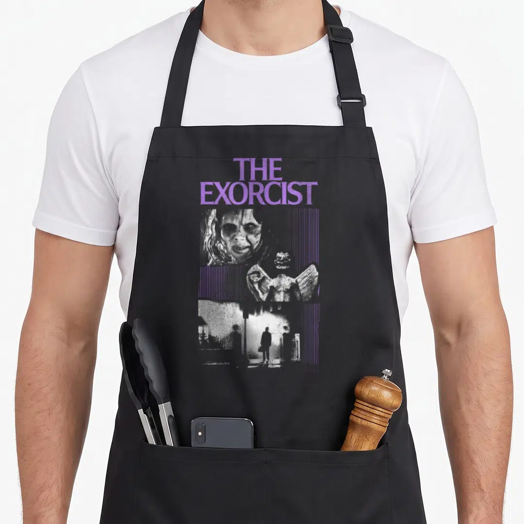 Exorcist novelty apron - THE EXORCIST purple text above three grey photos of a demon girl, winged creature, and man — shown on a black cotton canvas apron against a white background