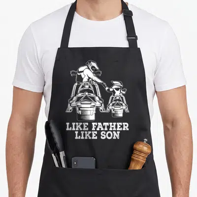Snowmobile father son apron - White father and son on snowmobiles above distressed like father like son text — shown on a black cotton canvas apron against a white background