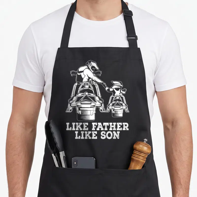 Snowmobile father son apron - White father and son on snowmobiles above distressed like father like son text — shown on a black cotton canvas apron against a white background