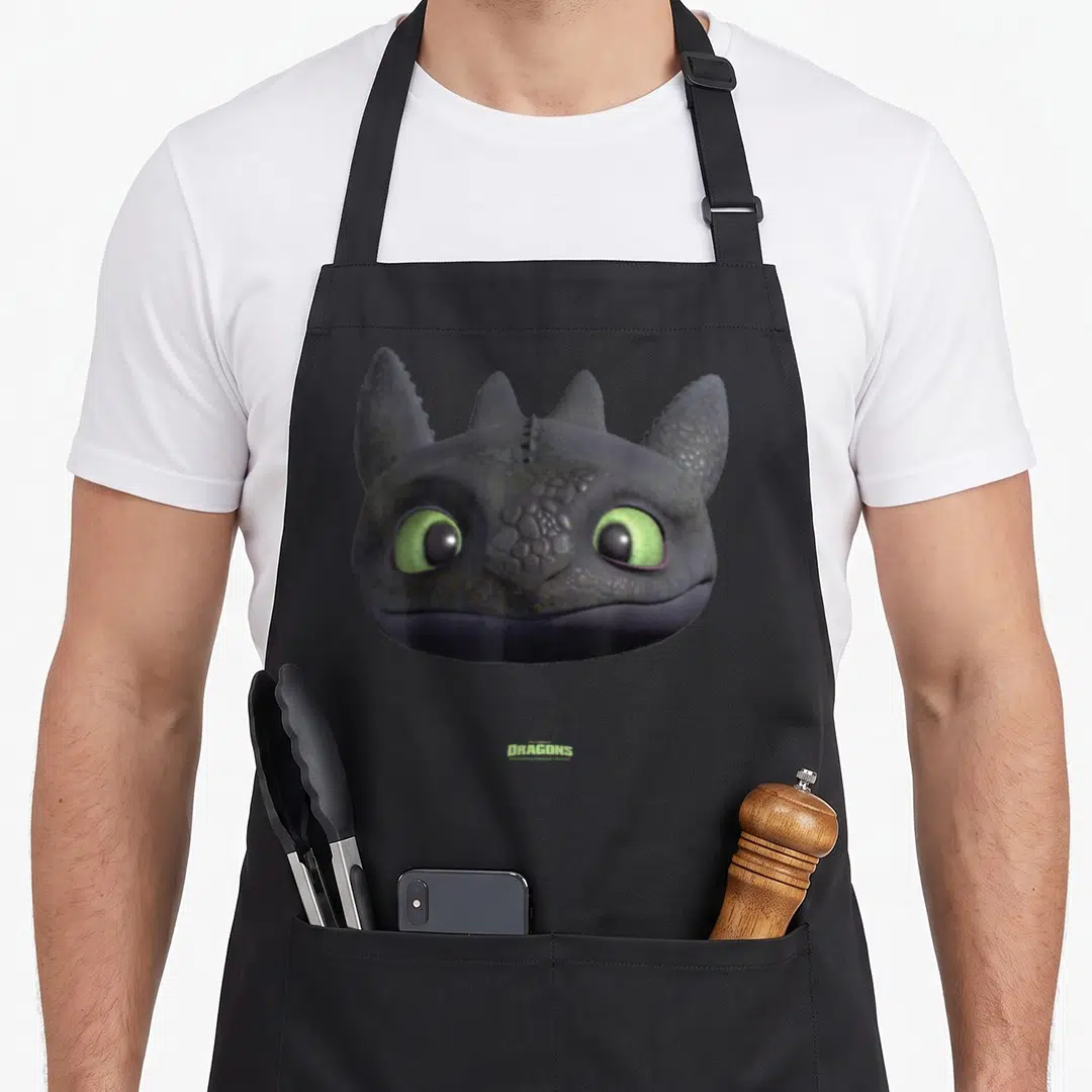 Toothless dragon apron - Grey dragon head with green eyes and green DRAGONS text centered at bottom — shown on a black cotton canvas apron against a white background