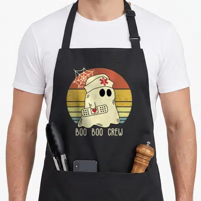 Boo nurse apron - Ghost nurse holding bandage with BOO BOO CREW text over retro sunset — shown on a black cotton canvas apron against a white background