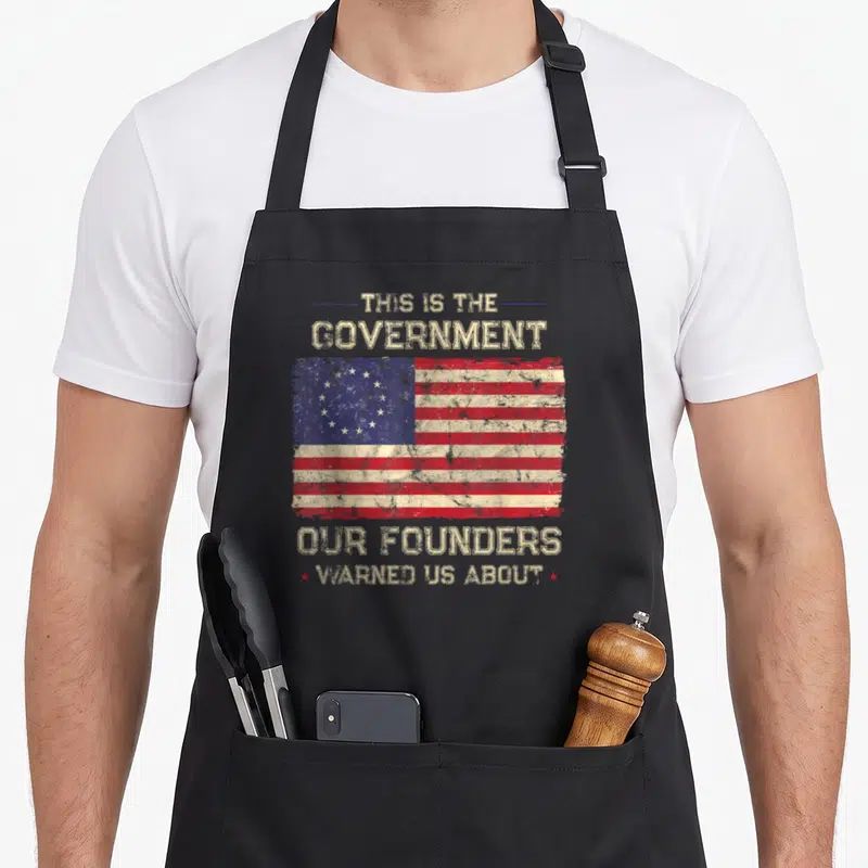 Government patriots apron - Distressed USA flag with text THIS IS THE GOVERNMENT OUR FOUNDERS WARNED US ABOUT in beige — shown on a black cotton canvas apron against a white background