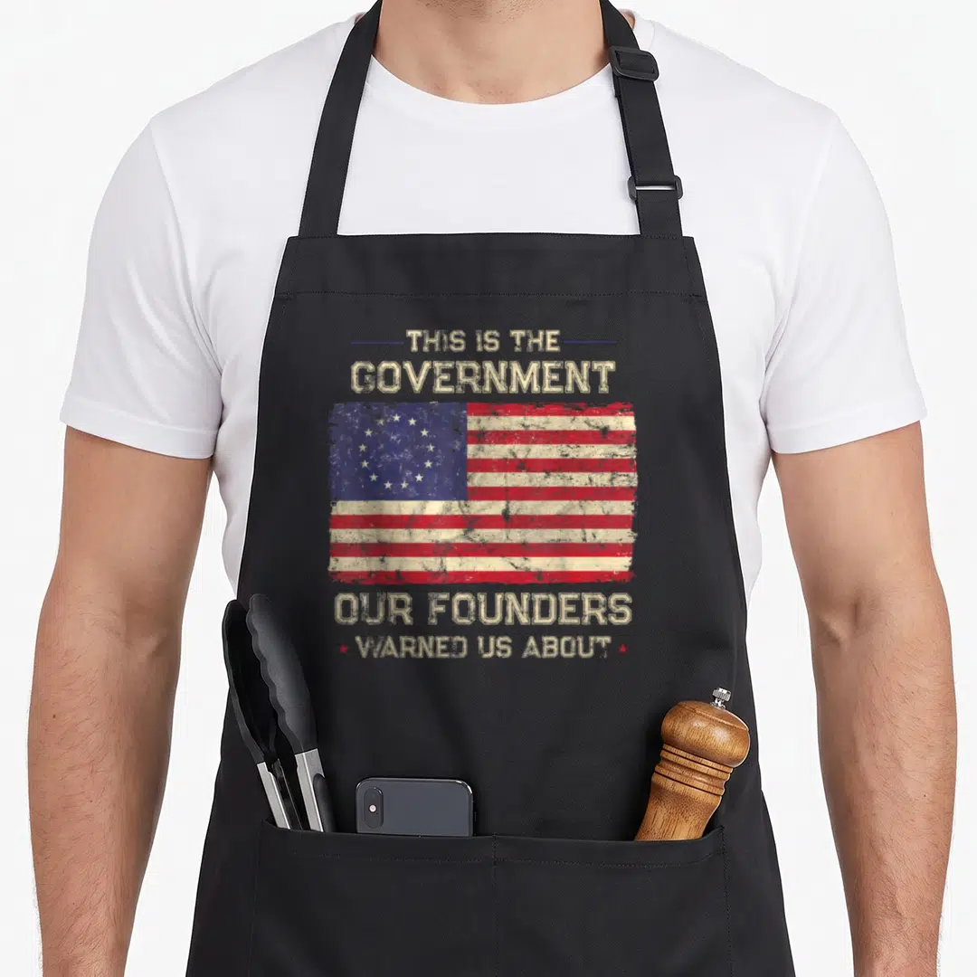 Government patriots apron - Distressed USA flag with text THIS IS THE GOVERNMENT OUR FOUNDERS WARNED US ABOUT in beige — shown on a black cotton canvas apron against a white background
