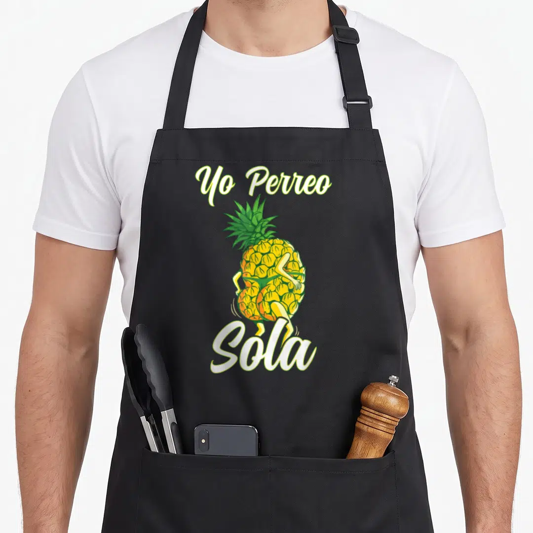 Perreo pineapple apron - Cartoon pineapple with limbs dancing between Yo Perreo and Sola script text — shown on a black cotton canvas apron against a white background