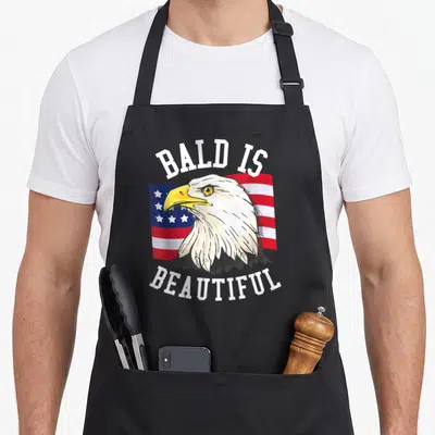 Bald beautiful eagles apron - Cartoon bald eagle head over waving flag with arched text BALD IS BEAUTIFUL in white — shown on a black cotton canvas apron against a white background
