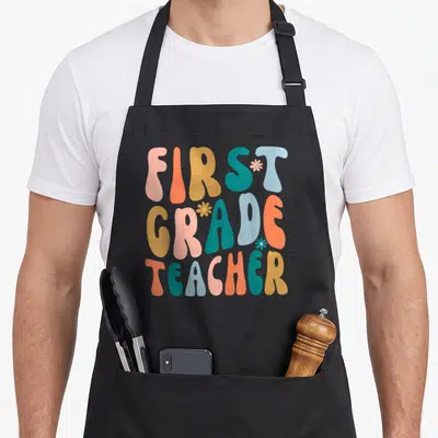 Grade groovy teacher apron - Retro text FIRST GRADE TEACHER in pink orange yellow teal blue with three small flowers — shown on a black cotton canvas apron against a white background