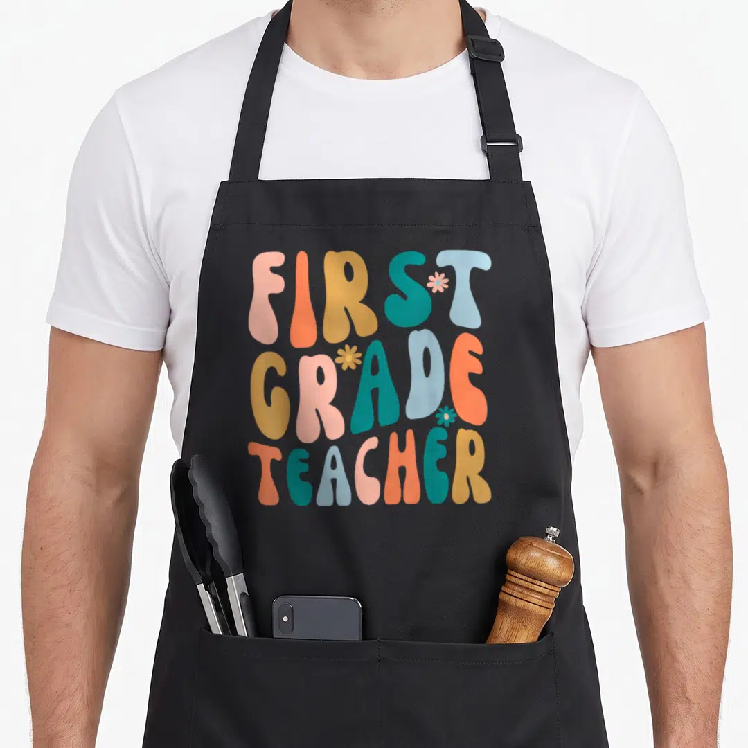 Grade groovy teacher apron - Retro text FIRST GRADE TEACHER in pink orange yellow teal blue with three small flowers — shown on a black cotton canvas apron against a white background