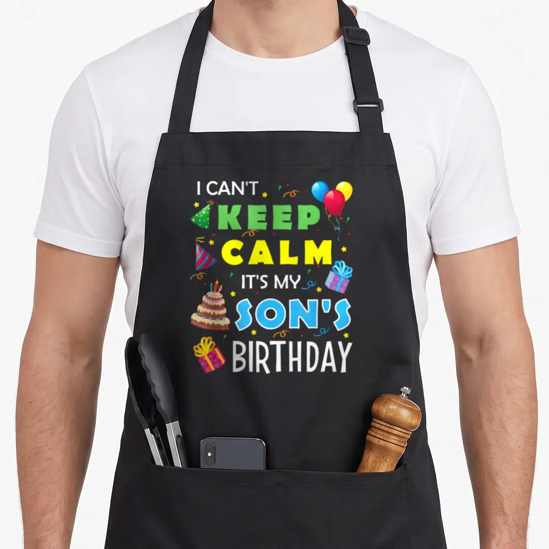 Son birthday apron - I Can't Keep Calm It's My Son's Birthday text with balloons, cake, and gifts in cartoon style — shown on a black cotton canvas apron against a white background