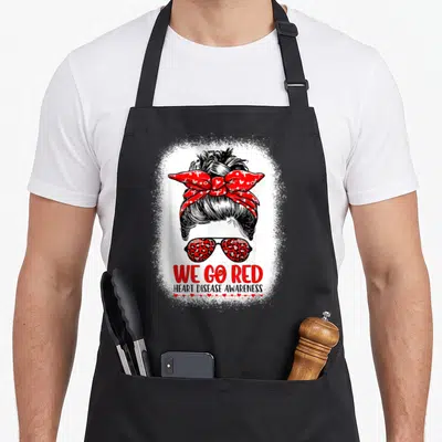 Disease messy aprons - Woman bun leopard sunglasses We Go Red text hearts — shown on a black cotton canvas apron against a white background