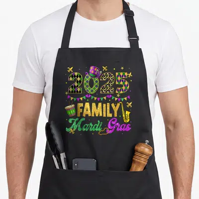 Family fat chef apron - 2025 FAMILY Mardi Gras design with drum saxophone beads and fleur-de-lis in purple green gold — shown on a black cotton canvas apron against a white background