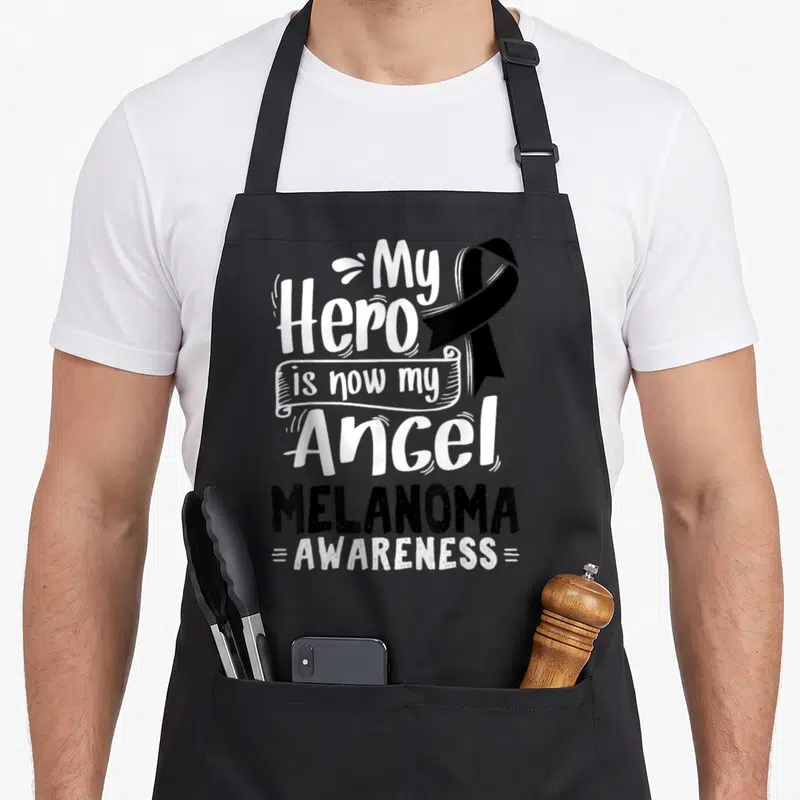 Malignant ribbon apron - White My Hero is now my Angel text with black ribbon and Melanoma Awareness lettering — shown on a black cotton canvas apron against a white background