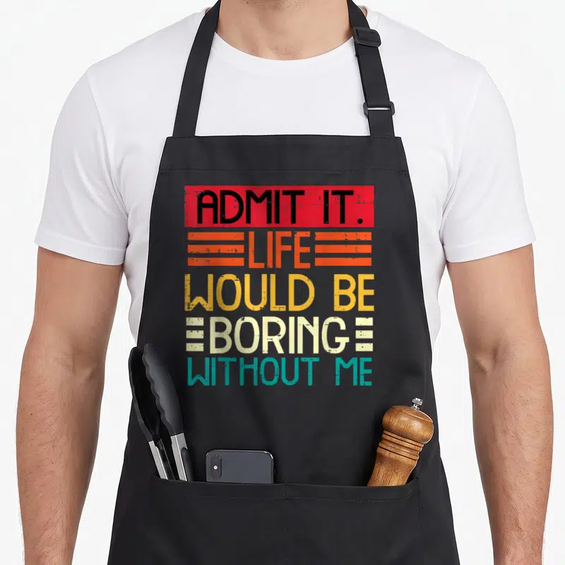 Funny boring novelty apron - Stacked colorful text ADMIT IT LIFE WOULD BE BORING WITHOUT ME on black background — shown on a black cotton canvas apron against a white background