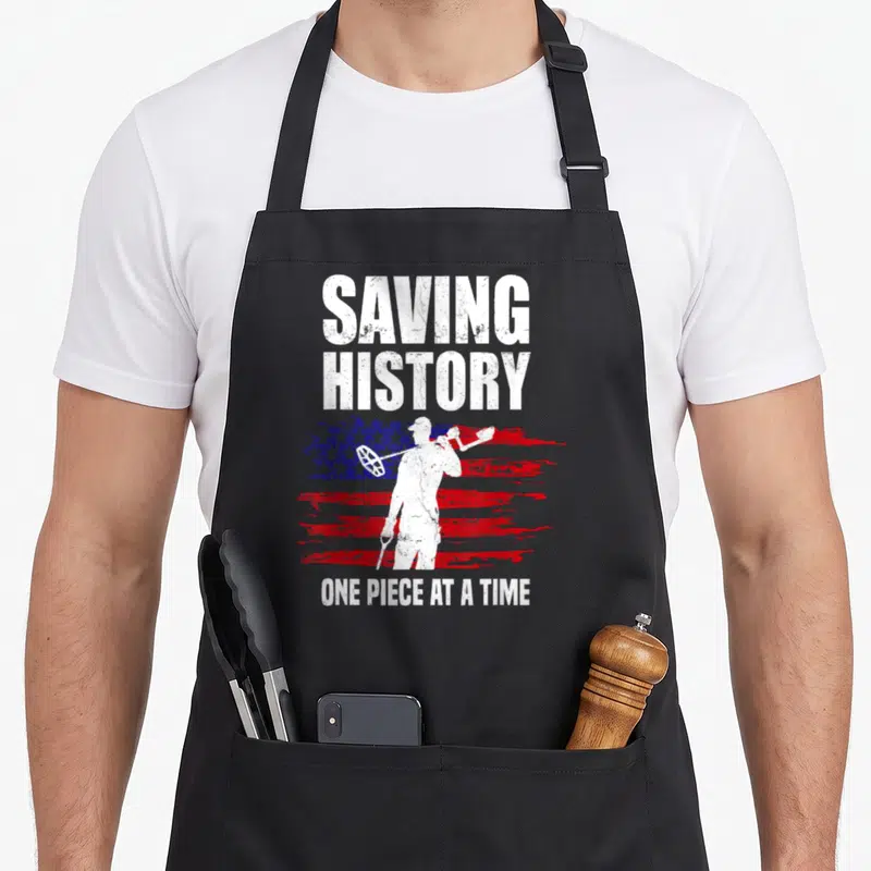 Saving american flag apron - White metal detectorist silhouette on distressed red white and blue flag with text — shown on a black cotton canvas apron against a white background