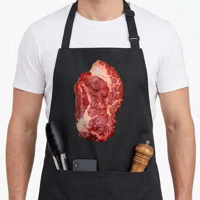Raw cow apron - Realistic raw red and white steak photo centered on black fabric — shown on a black cotton canvas apron against a white background