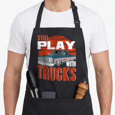 Play truck apron - Retro blue pickup truck with red sun and STILL PLAY WITH TRUCKS text — shown on a black cotton canvas apron against a white background