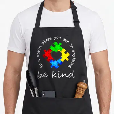 Anything young at heart apron - Four colorful puzzle pieces forming a heart with circular text reading be kind — shown on a black cotton canvas apron against a white background