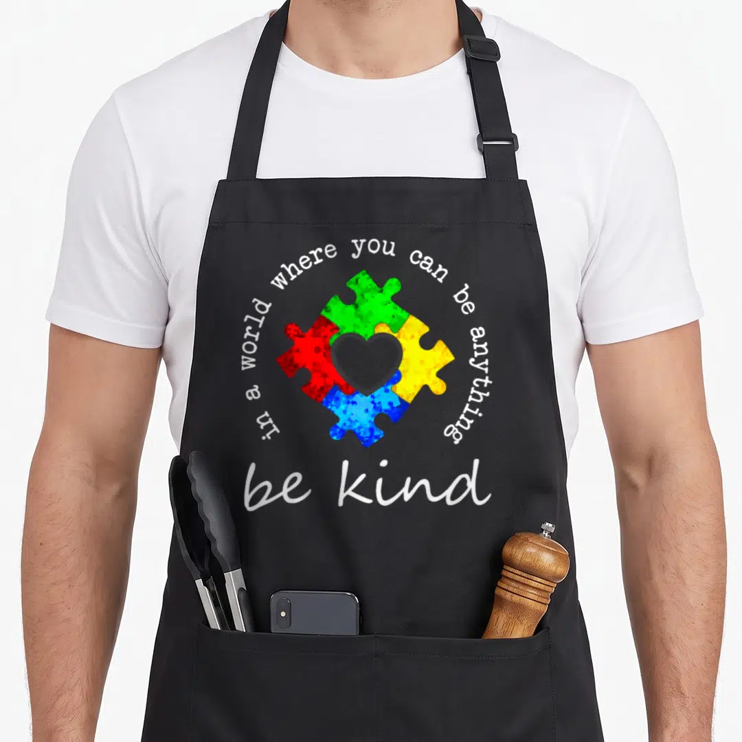 Anything young at heart apron - Four colorful puzzle pieces forming a heart with circular text reading be kind — shown on a black cotton canvas apron against a white background