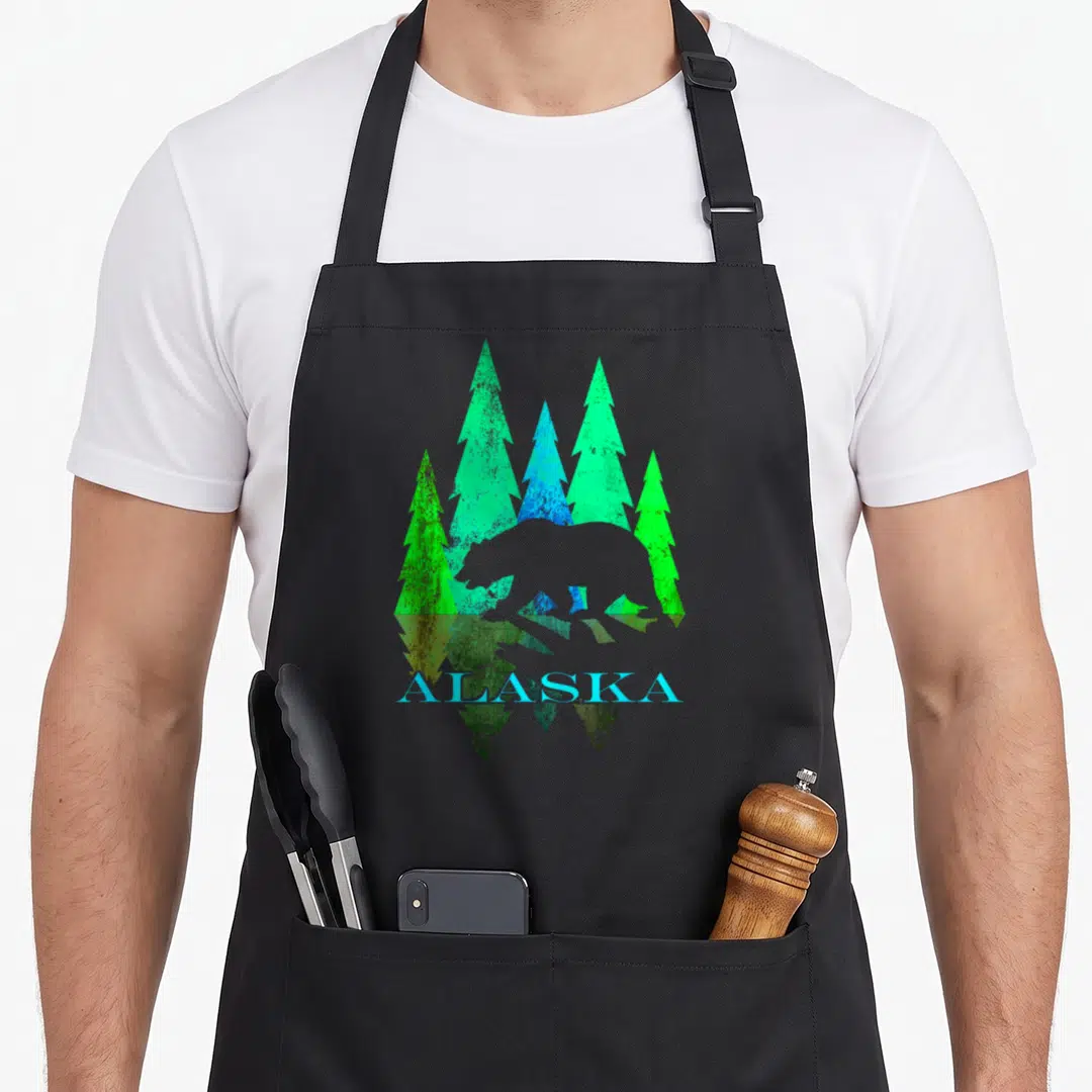 Alaska alaskan bear apron - Minimalist bear silhouette over green and blue pine trees with ALASKA text and vertical — shown on a black cotton canvas apron against a white background