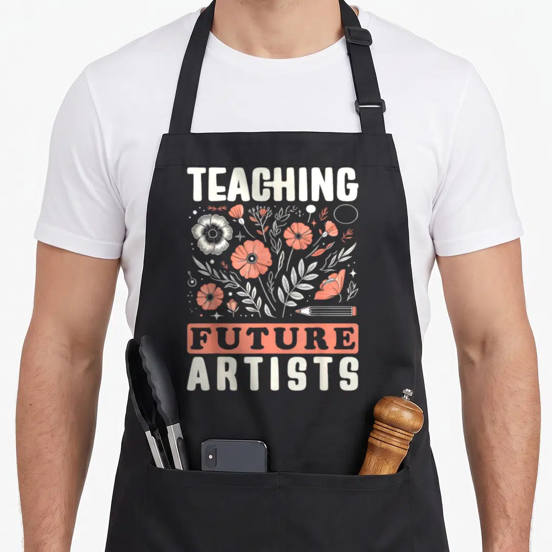 Future artist teacher apron - White and orange flowers surrounding a pencil with TEACHING FUTURE ARTISTS text — shown on a black cotton canvas apron against a white background