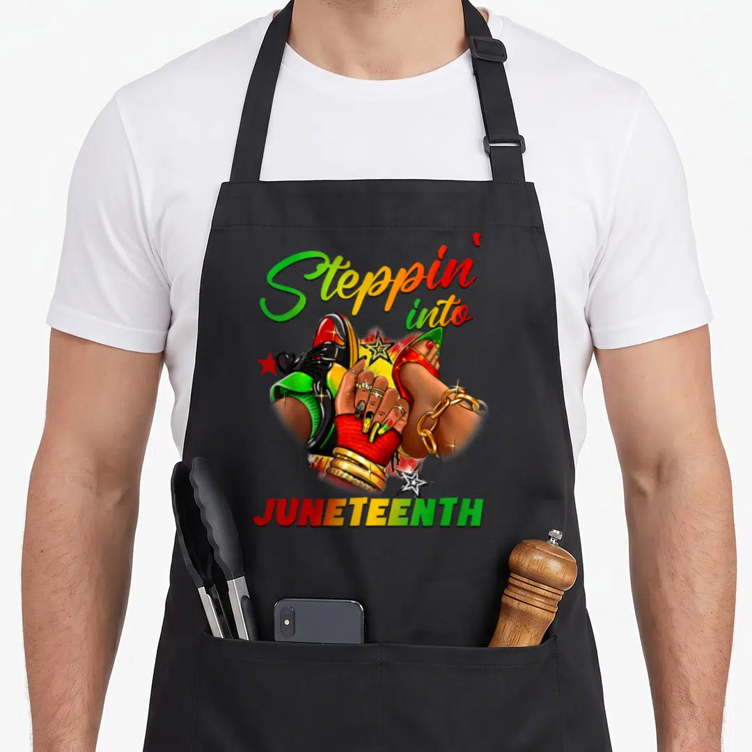 Juneteenth stepping apron - Arched Steppin' into JUNETEENTH text above cartoon feet, hand, and stars in green, yellow — shown on a black cotton canvas apron against a white background
