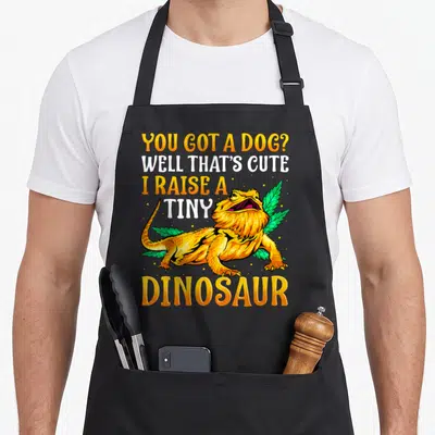 Pet dragon apron - Bearded dragon illustration with stacked text YOU GOT A DOG? WELL THAT'S CUTE I RAISE A TINY — shown on a black cotton canvas apron against a white background