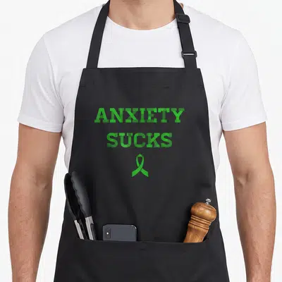 Suck anxiety awareness apron - Green ANXIETY SUCKS text stacked above a green awareness ribbon — shown on a black cotton canvas apron against a white background
