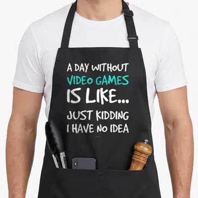 Funny video game apron - Stacked white and teal text reading A DAY WITHOUT VIDEO GAMES IS LIKE JUST KIDDING I HAVE NO — shown on a black cotton canvas apron against a white background