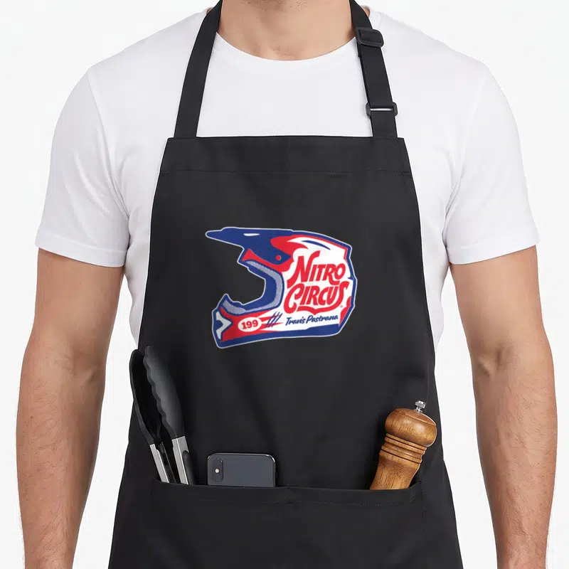 Nitro circus apron - Blue red and white helmet graphic with Nitro Circus text and Travis Pastrana name — shown on a black cotton canvas apron against a white background
