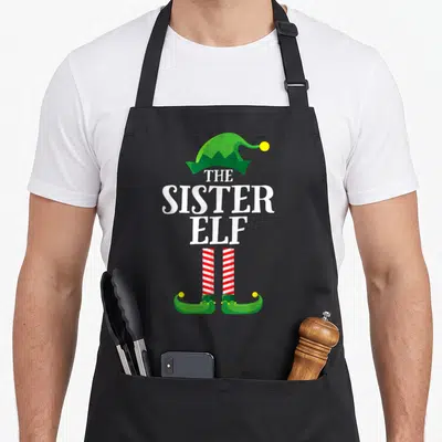 Sister elf apron - Stacked text THE SISTER ELF between green elf hat and red striped legs with yellow pom-poms — shown on a black cotton canvas apron against a white background