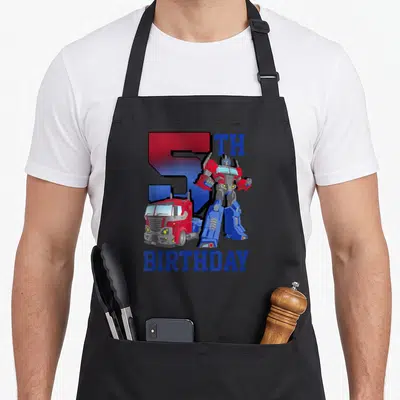 Gradient birthday apron - Optimus Prime robot and truck with large number 5 and birthday text in red and blue — shown on a black cotton canvas apron against a white background