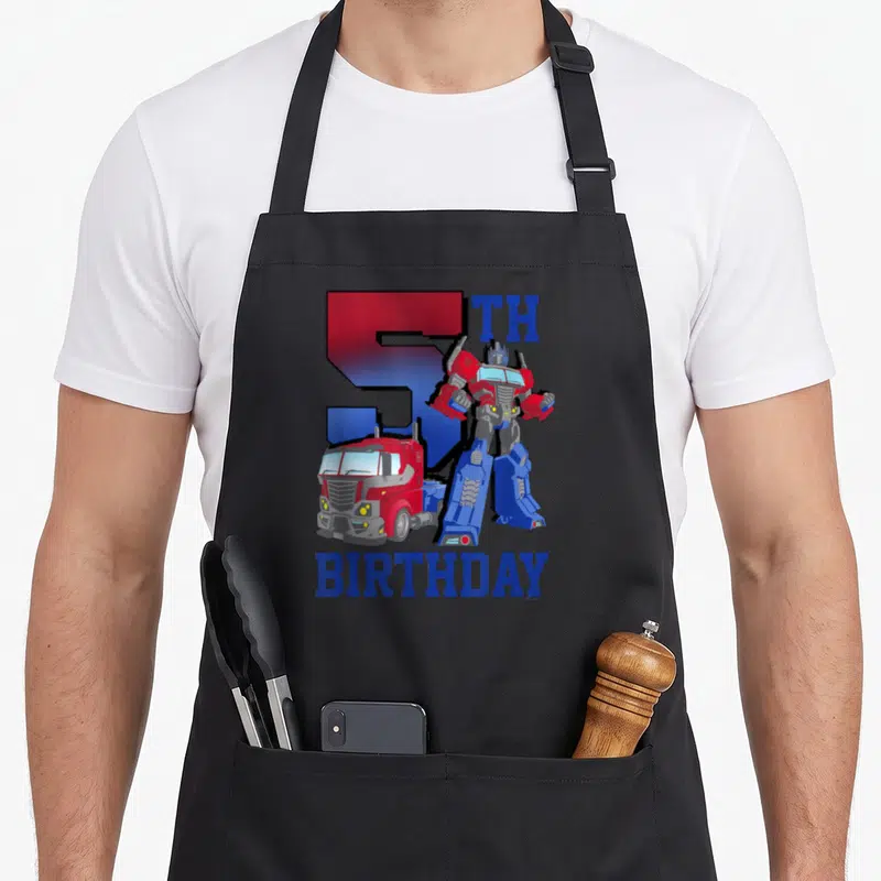 Gradient birthday apron - Optimus Prime robot and truck with large number 5 and birthday text in red and blue — shown on a black cotton canvas apron against a white background