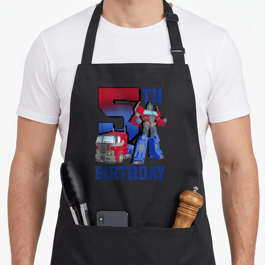 Gradient birthday apron - Optimus Prime robot and truck with large number 5 and birthday text in red and blue — shown on a black cotton canvas apron against a white background