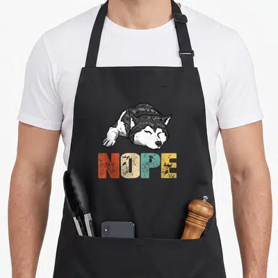 Nope husky apron - Cartoon lying husky above multicolor NOPE text in red beige blue yellow grey white — shown on a black cotton canvas apron against a white background