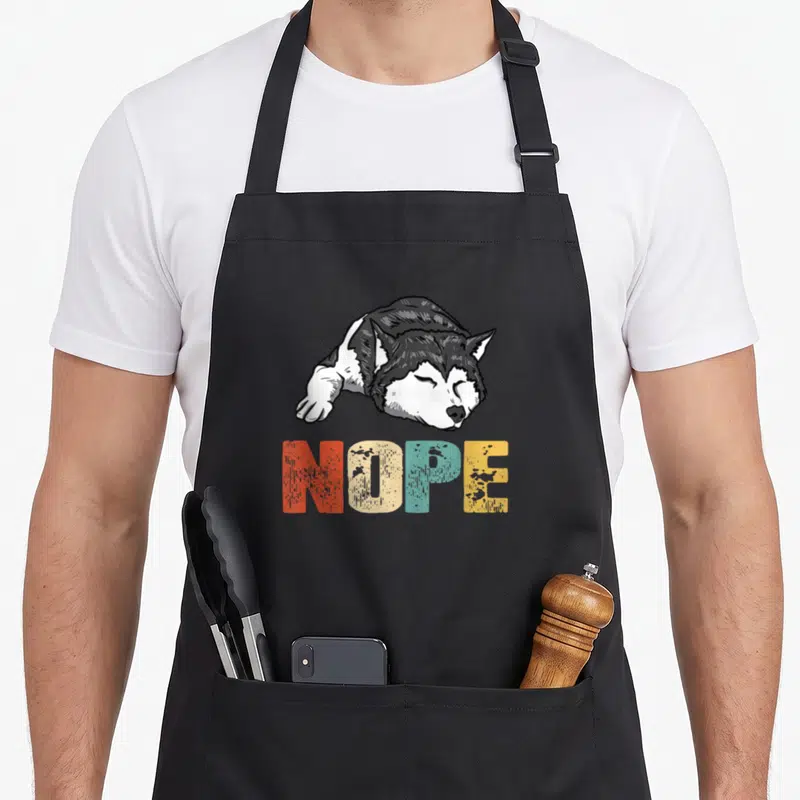Nope husky apron - Cartoon lying husky above multicolor NOPE text in red beige blue yellow grey white — shown on a black cotton canvas apron against a white background