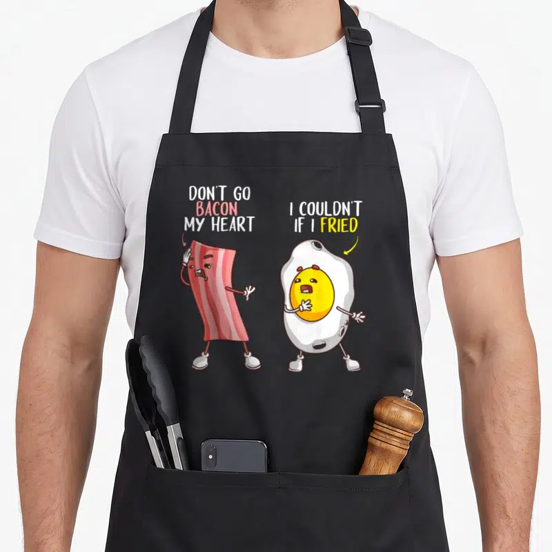 Egg bacon apron - Cartoon bacon and fried egg with text Don't Go Bacon My Heart I Couldn't If I Fried — shown on a black cotton canvas apron against a white background