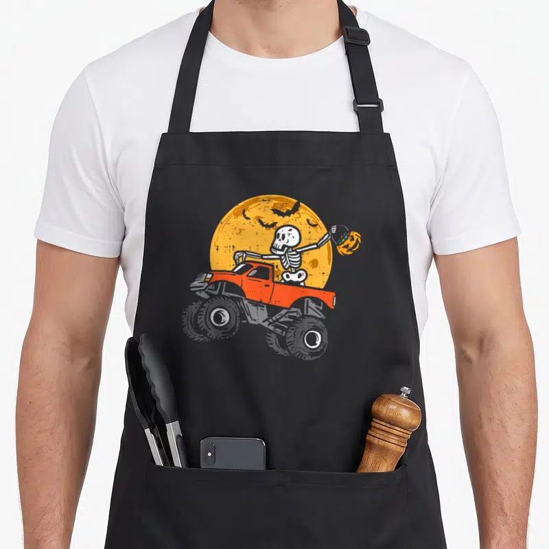 Skeleton cotton candy apron - Cartoon skeleton in red truck holding pumpkin under yellow moon with flying bats — shown on a black cotton canvas apron against a white background