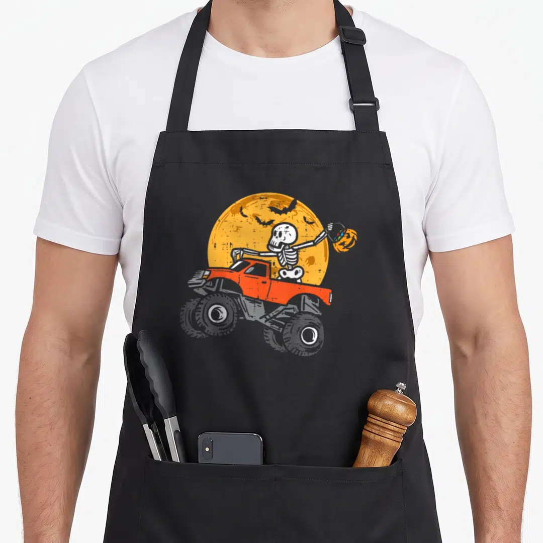 Skeleton cotton candy apron - Cartoon skeleton in red truck holding pumpkin under yellow moon with flying bats — shown on a black cotton canvas apron against a white background