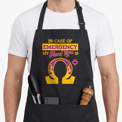 Psi blood apron - Stacked text reading IN CASE OF EMERGENCY above a patterned gold and purple omega symbol with a plus — shown on a black cotton canvas apron against a white background
