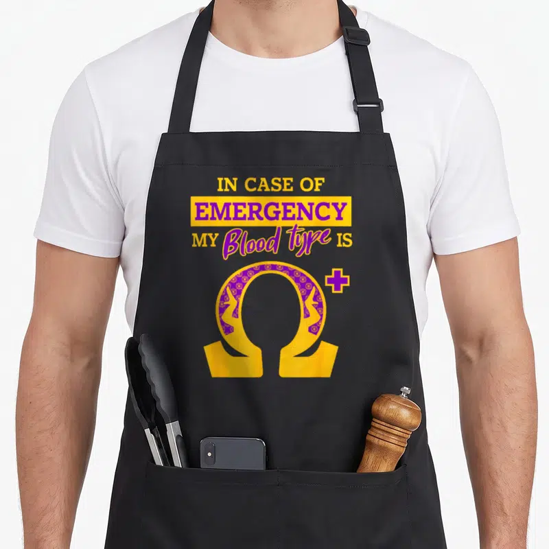 Psi blood apron - Stacked text reading IN CASE OF EMERGENCY above a patterned gold and purple omega symbol with a plus — shown on a black cotton canvas apron against a white background