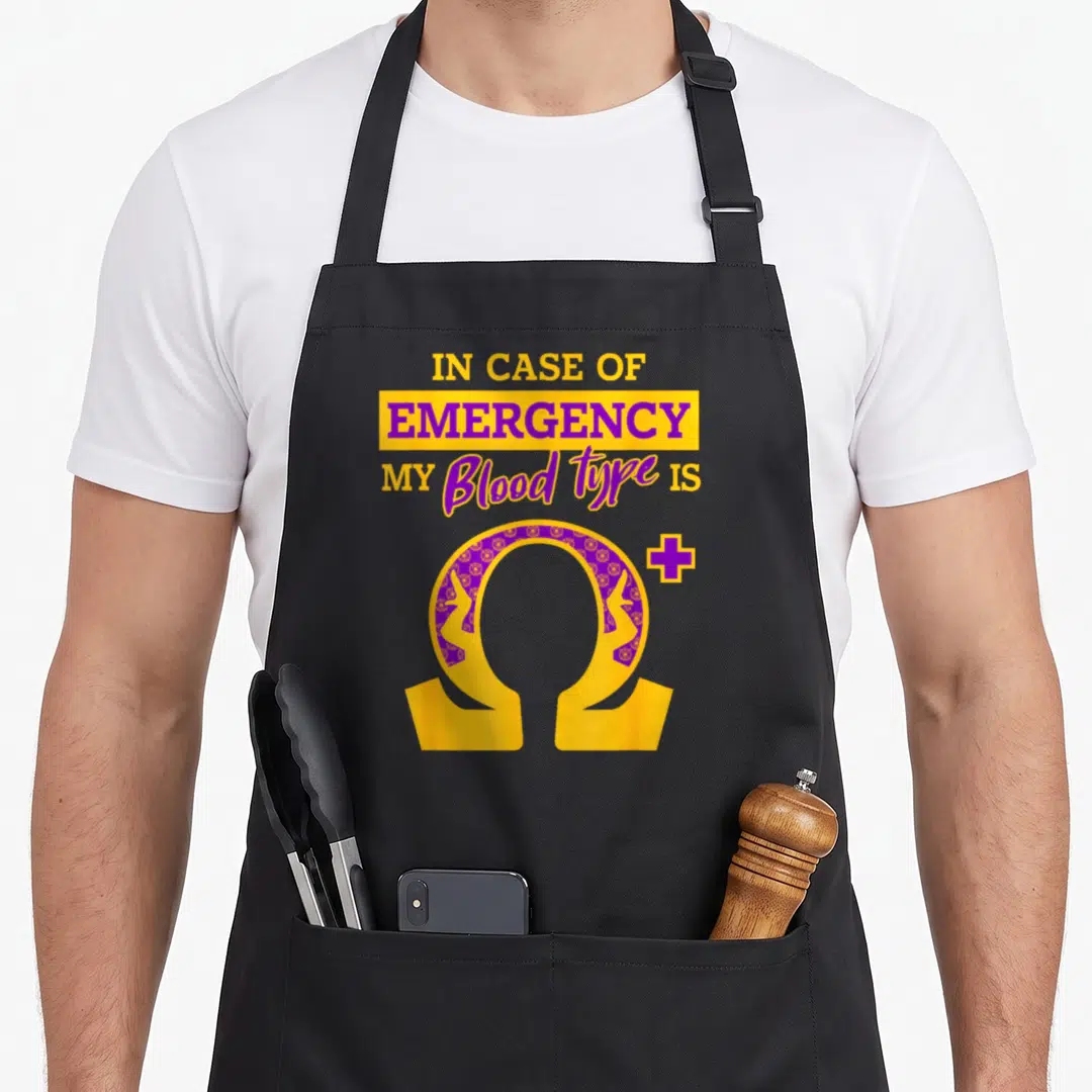 Psi blood apron - Stacked text reading IN CASE OF EMERGENCY above a patterned gold and purple omega symbol with a plus — shown on a black cotton canvas apron against a white background