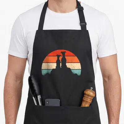 Whippet sunset dog apron - Two black dog silhouettes sitting in grass against a striped retro sun with red, orange — shown on a black cotton canvas apron against a white background
